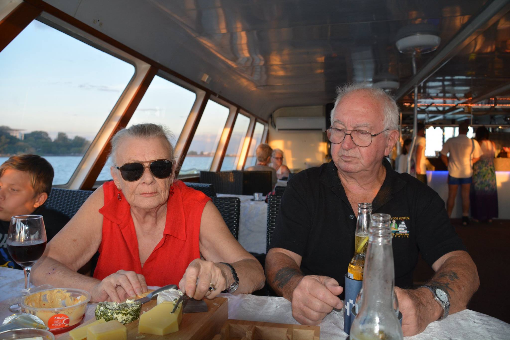 Rhonda and Barry sitting side by side at a table on a boat at sunset, with a cheese platter and drinks in front of them