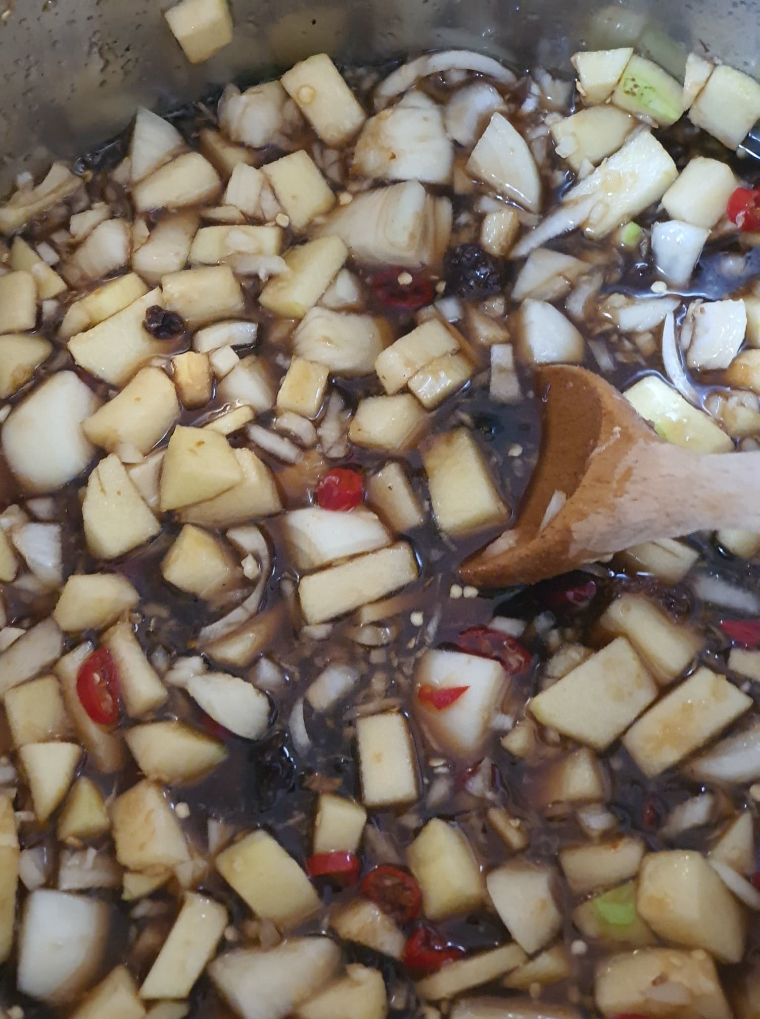 Diced apple and onion, raisins and sliced red chilli in the dark liquid, being stirred with a wooden spoon